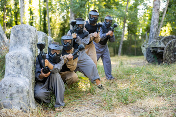 Fototapeta premium Group of men and women in protective clothes and helmets shooting with paintball markers while hiding behind cover. Teams fighting on battlefield outdoors.