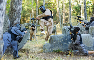 Fototapeta premium Skillful paintball team in protective outwear and helmets attacking hostile positions, running and jumping over enemy covers.