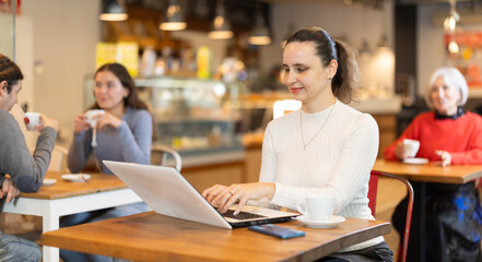 Portrait of focused female surfing Internet on notebook while drinking tea or coffee in cafeteria