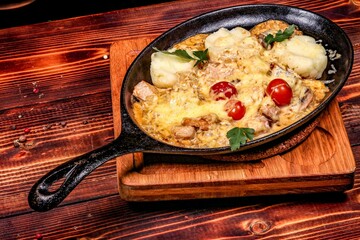 Cozy baked meat with cheese and potatoes in skillet on board, angled view on wooden background.