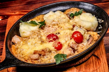Hot baked meat with cheese sauce and potatoes in cast iron skillet, close-up angled view on wood.