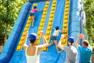 Two young women competing in climbing with wood poles on tall inflatable slide on adults bouncy...