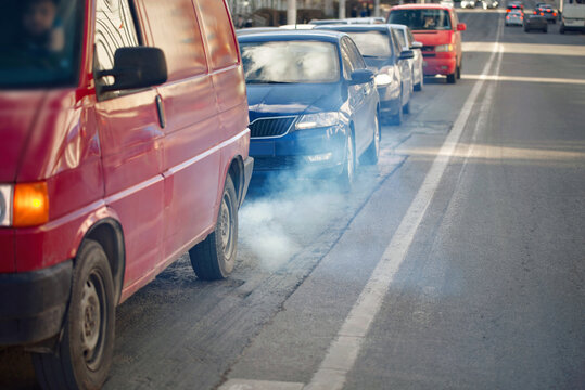 Smoke rises from van exhaust in line of cars, impact of vehicle emissions and air pollution. City traffic emits vehicle exhaust into the air, harmful emissions. Tailpipe smoke. Selective focus.