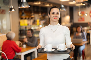 Woman visitor in self-service cafeteria carries tray with two cups of coffee. Customer has purchased freshly brewed coffee for herself and companion