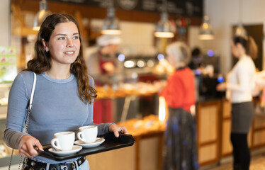 Girl with two cups of coffee on tray goes to table in cafe, having coffee break with friends, colleagues