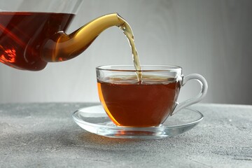 Pouring freshly brewed tea from teapot into cup at grey table, closeup