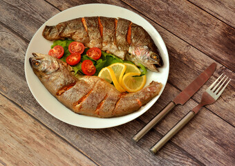 A plate with two whole baked trouts with spinach, lemon and cherry tomatoes on a wooden table.