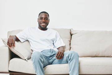 Fototapeta premium Smiling young Black man relaxing on a beige couch, wearing a casual white t shirt and light blue jeans, exuding a feeling of comfort and happiness