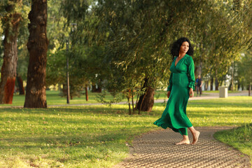 Fototapeta premium Young barefoot curly dark haired woman in green dress walking in the park