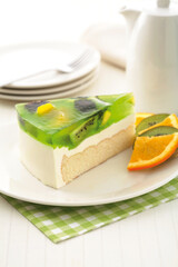 Close-up of a slice of kiwi cheesecake with a clear green jelly topping, kiwi slices, and orange wedges on a white plate.