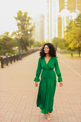Pretty young curly dark haired woman in green dress walking in the street early in the morning full...