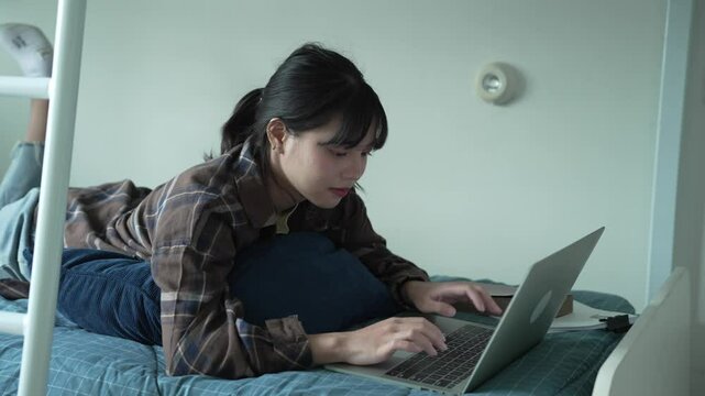 Asian girl prone on bed and typing on laptop