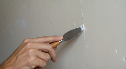 Hand applying putty to wall crack with metal spatula. Close-up of repair technique on white surface. Home maintenance and drywall restoration solutions