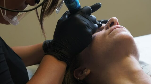 Slow motion of esthetician uses a blue tool to microblade a client&rsquo;s eyebrow. with black gloves, tilted head, and soft lighting highlighting precision and calm during this beauty tatooing procedure.