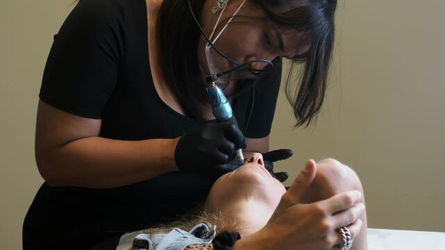Client rests with hand on forehead while esthetician uses a blue tool to perform microblading, both focused in a detailed and precise moment &mdash; slow motion mid-range shot from frontal angle.