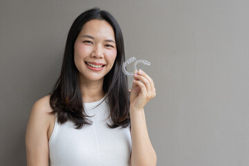 close up young asian beautiful woman smiling with hand holding dental aligner retainer (invisible) on grey background at dental clinic for beautiful teeth treatment course concept