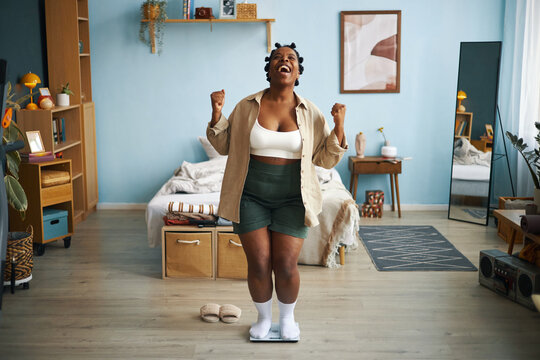 Black woman standing on scale in bedroom and cheering, celebrating weight loss success with raised arms, surrounded by cozy furniture and modern decor