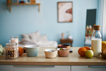 Healthy breakfast spread including fresh fruits, assorted bowls, glass carafe of milk on wooden countertop with blurred bedroom background