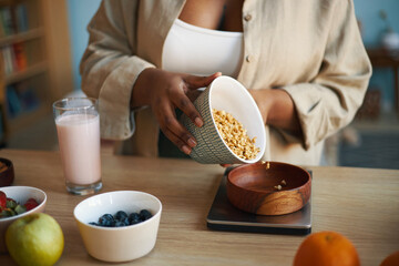Woman pouring granola into a bowl with a selection of fruits and a glass of milk. Scene set in a kitchen with various breakfast options