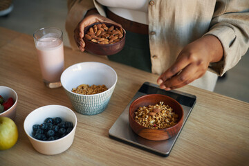 Close-up of hands preparing breakfast with granola, nuts, and fresh fruit in bowls with glass of...