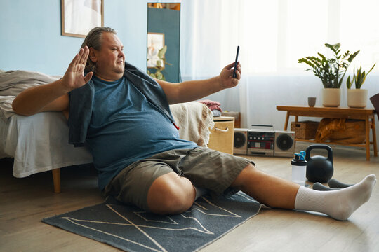 Caucasian man taking selfie while sitting on exercise mat in cozy bedroom with fitness equipment and bright natural light filling room - Powered by Adobe