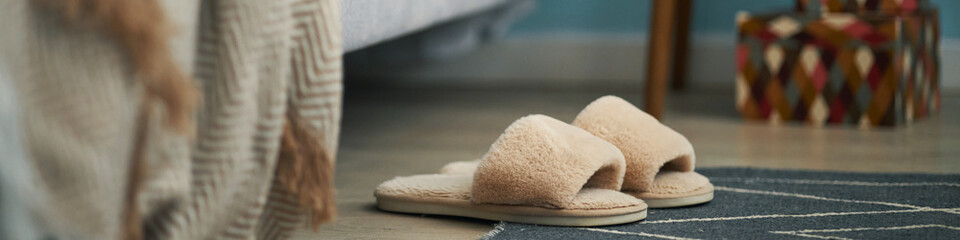 Fuzzy beige slippers placed on a blue floor rug next to an unmade bed, with part of a colorful box in background creating a cozy atmosphere