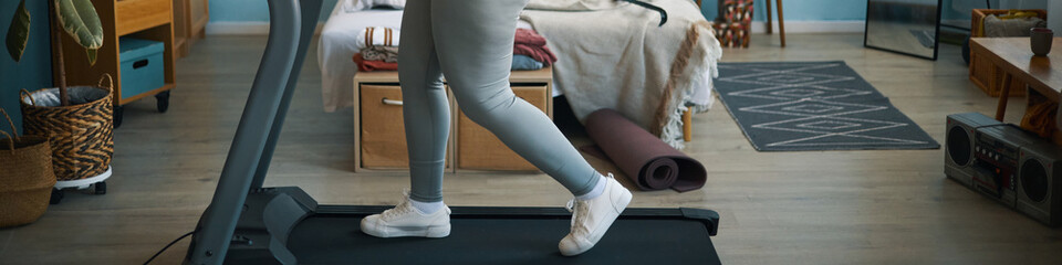 Woman in gray leggings and white sneakers exercising on treadmill in cozy home with wooden elements...