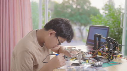 Asian teenage boy soldering electronic components of a robotics project at home, showing hands-on STEM education, electronics skills, and focus in home-based learning for young engineers.