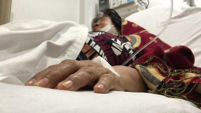 mature woman lies in a hospital bed with a dextrose IV attached to her right hand.