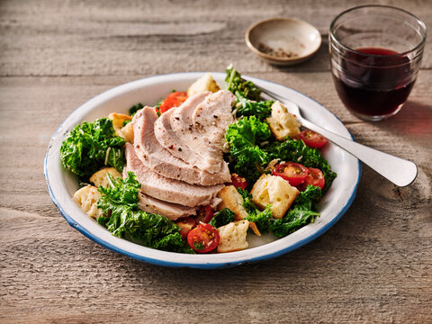 A vibrant turkey kale salad topped with sliced turkey breast, cherry tomatoes, and croutons is presented on a rustic plate. A glass of red beverage adds to the inviting atmosphere