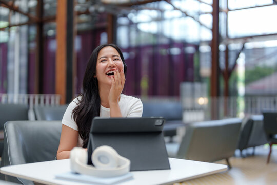 Asian freelancer is sitting at a table in a coworking space, laughing while having a video call on her tablet, enjoying a moment of levity during her workday