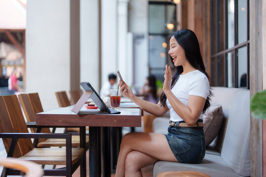 Happy young asian woman is waving and making a video call using a smartphone in a cafe, with a tablet and a glass of iced coffee on the table