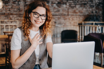 A young woman with curly red hair waves at her laptop screen during a virtual call in a cafe. Remote connection, online community, digital interaction, friendly virtual presence...