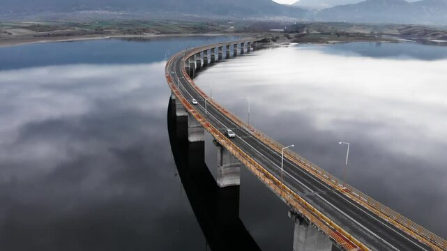 wide aerial View of High Bridge of Servia at lake polyfytos Kozani greece while tracking following cars , we see the reflection of the clouds at the lake and the bridge at the middle of the frame