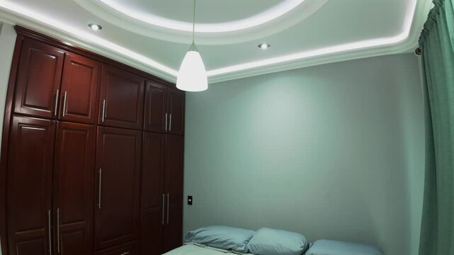 Tilt down from large circular ceiling cove with recessed lighting to a small bedroom with a bed, pale blue linens, and wooden built-in wardrobes