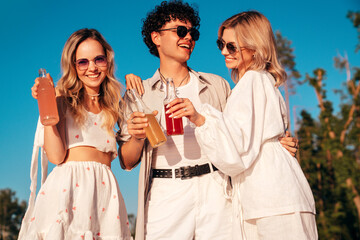 Young stylish friends posing at the seaside. Fashion man and two cute female dressed in casual summer clothes. Smiling models having fun. Cheerful women and guy outdoors, Hold and drink lemonade