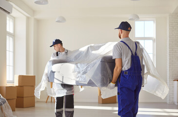 Moving day and loading service concept. Two male moving service workers in coveralls carrying sofa,...