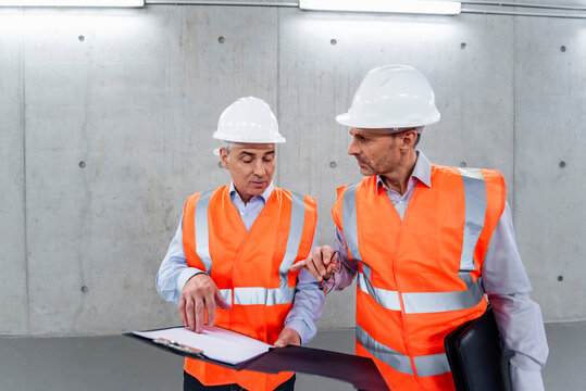 Two engineers in safety vests and hard hats discussing plans in a storage hall