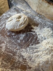A single piece of dough sits on a floured wooden surface, ready for baking, with a rustic loaf and scattered flour nearby in soft lighting.