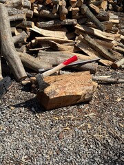 A splitting maul embedded in a log, surrounded by chopped and unchopped firewood on gravel under...