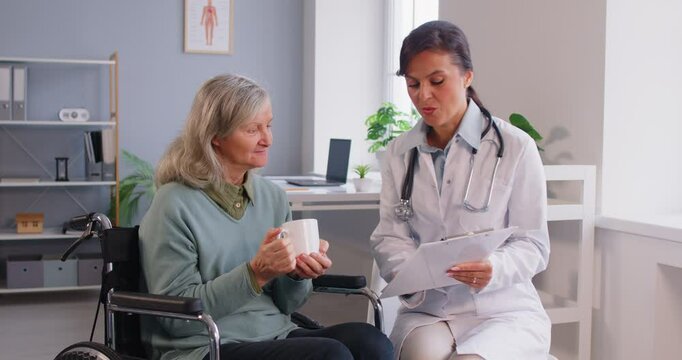 Elderly care. Medical professional is engaging in discussion with elderly woman seated in wheelchair. Elderly woman holds cup of hot drink and listens attentively to doctor who shows her clipboard.