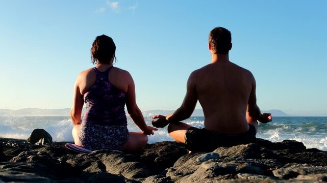 Couple meditate at sunrise on coastal rocks after swim facing crashing waves