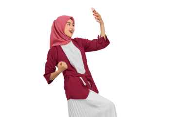 Excited young Asian Muslim woman in hijab celebrating success with smartphone and showing victory gesture isolated on transparent background