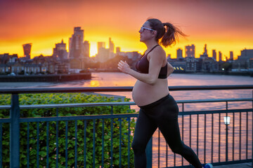 A pregnant woman in sports clothing running for exercise in front of an urban skyline during a fiery sunset