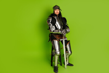 Medieval knight in armor with sword sitting on stool against vibrant green background