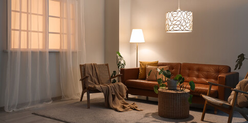 Interior of living room with brown sofa, armchairs, table and glowing lamps in evening
