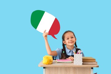 Happy schoolgirl with speech bubble sitting at desk against light blue background. Studying of...
