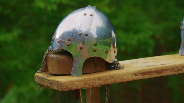 Close-up shot of a shiny medieval helmet crafted from polished metal, sitting atop a wooden platform. The helmet features a rounded top and is adorned with chainmail coif