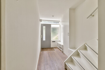 A spacious, well-lit hallway featuring a simple design with white walls and wooden flooring, complemented by a sleek staircase.