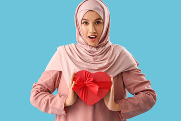 Beautiful surprised Muslim woman with gift box for Valentine's Day on blue background